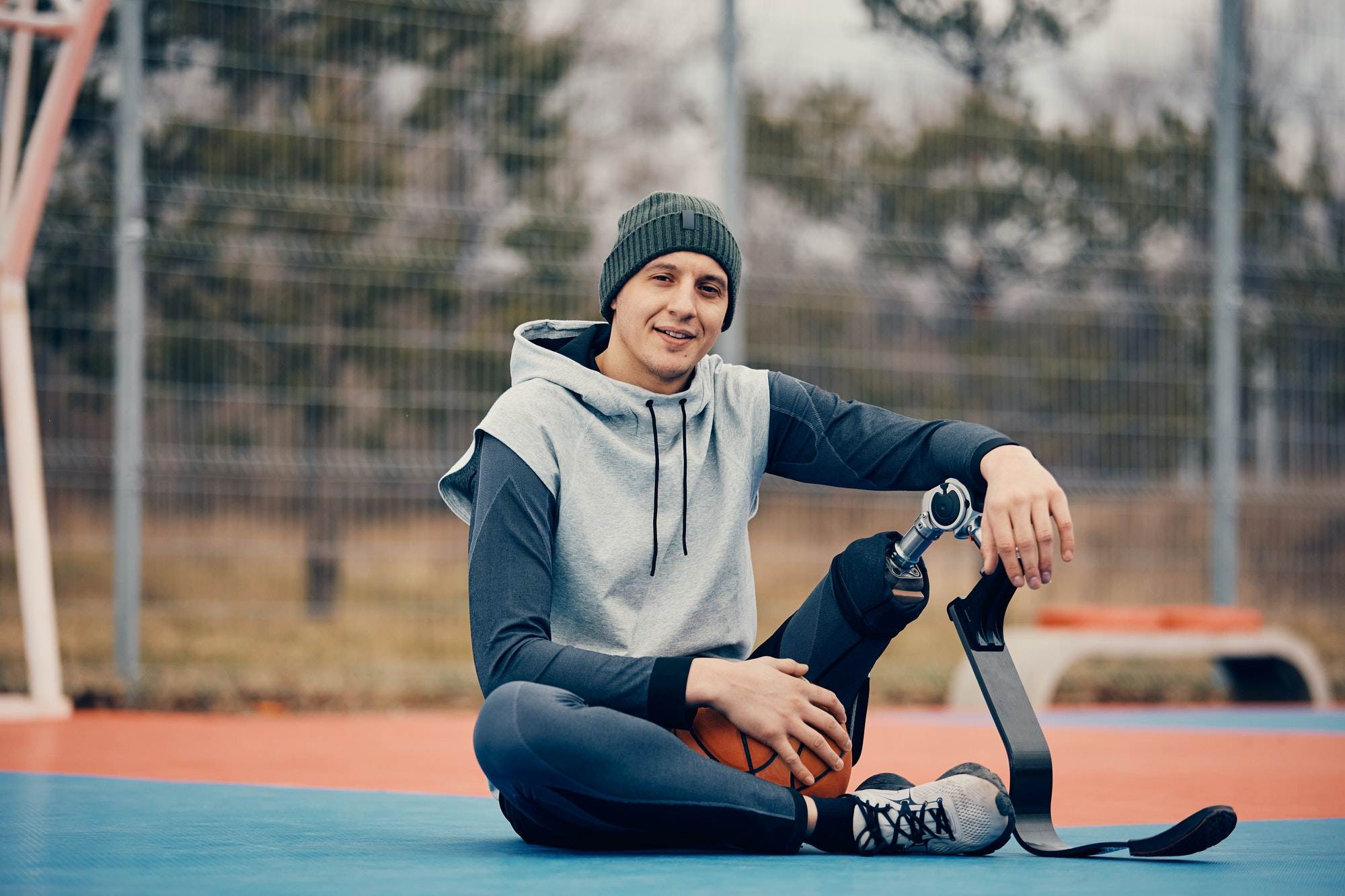 Young athlete with prosthetic leg relaxing on outdoor basketball court and looking at camera.